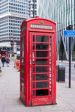 London, United Kingdom - 12 May 2025: View of a classic red telephone box on a city sidewalk with modern architecture and pedestrians in the background.
