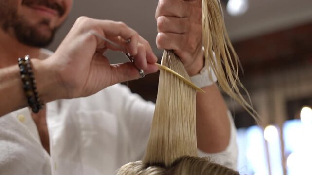 Skilled male hairdresser trimming a woman's wet blonde hair. Stylist using scissors and a comb for a precision haircut in a modern spa