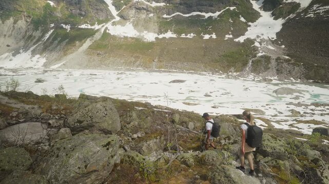 Static shot of a hiking couple crossing massive boulders above a glacier valley with visible moraine and rugged alpine wilderness scenery at Austerdalsbreen Glacier Norway