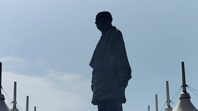 Zoom-in on Statue of Unity full silhouette rising above tensile canopy mast structures against soft blue sky. For India heritage docs, patriotic content, and landmark sequences.