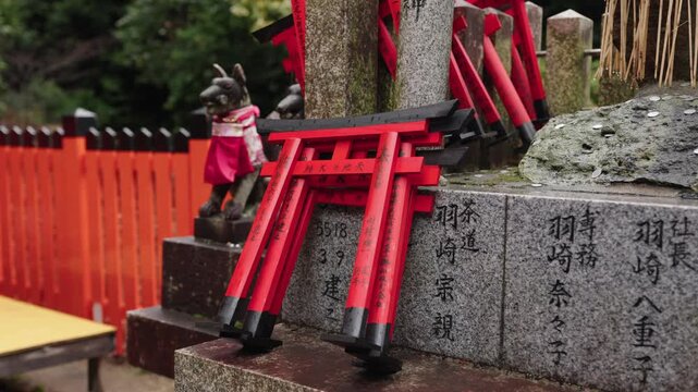 Small red votive torii gates stacked on carved stone with kanji inscriptions beside a kitsune statue at Fushimi Inari Kyoto