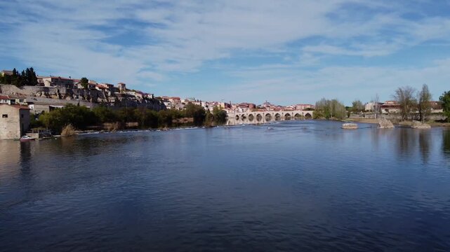 4K aerial drone video flying at a very low altitude over the calm and tranquil waters of the Duero River in Zamora, Spain. The smooth low-angle shot captures the historic city, lush trees, and the ico
