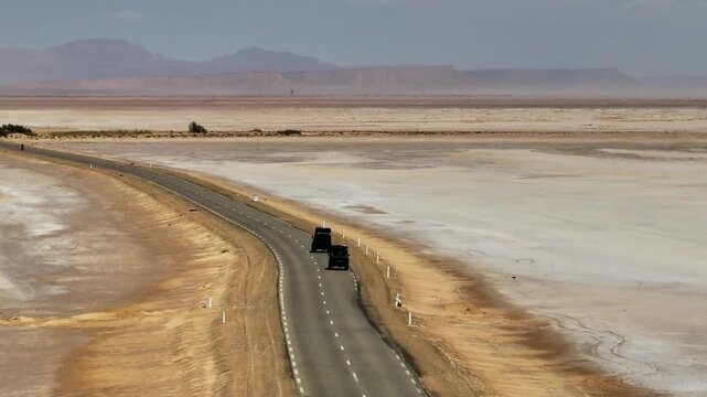 Off-road vehicles on the asphalt road crossing Chott el Djerid salt lake. salt flats and desert horizon in southern Tunisia aerial drone