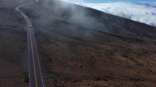 Drone pullback shot from the summit road of Haleakala volcano on Maui Hawaii with dramatic clouds rolling over the volcanic landscape above the clouds