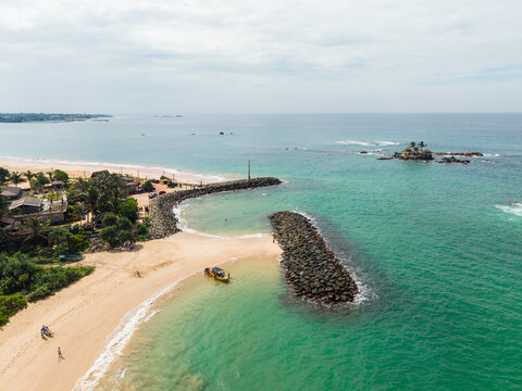 Aerial view of turquoise ocean waves hitting a sandy beach with stone breakwaters and a small boat in Peraliya, Southern Province, Sri Lanka.