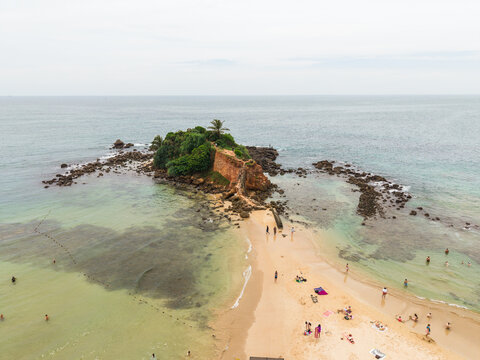 Aerial view of Parrot Rock, a small rocky outcrop with lush vegetation, connected to a sandy beach with people swimming and sunbathing in Mirissa, Southern Province, Sri Lanka.