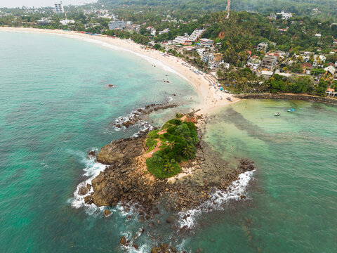 Aerial view of Parrot Rock island connected to the sandy beach by a narrow sandbar with turquoise ocean water and coastal buildings Mirissa, Southern Province, Sri Lanka.