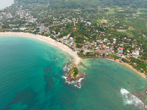 Aerial view of Parrot Rock and the sandy shoreline of Mirissa Beach with turquoise ocean water and lush tropical vegetation in Mirissa, Southern Province, Sri Lanka.