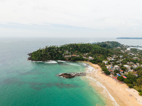 Aerial view of the tropical coastline of Mirissa Beach featuring Parrot Rock, turquoise waters, and lush palm trees in Mirissa, Southern Province, Sri Lanka.