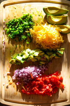 Fresh chopped and grated taco ingredients including tomato, onion, avocado, jalapeno pepper, and cheddar cheese on a cutting board ready to be put in a shell and eaten for dinner. 