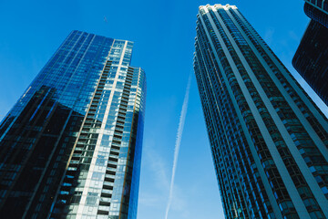Fototapeta premium View of tall buildings reaching the sky with a clear blue background and a trail in the air showing a plane flying high above the city