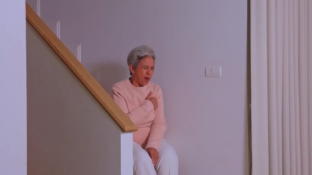Worried elderly woman experiencing sudden severe chest pain on wooden staircase inside modern home feeling intense distress during acute senior medical emergency heart attack illness