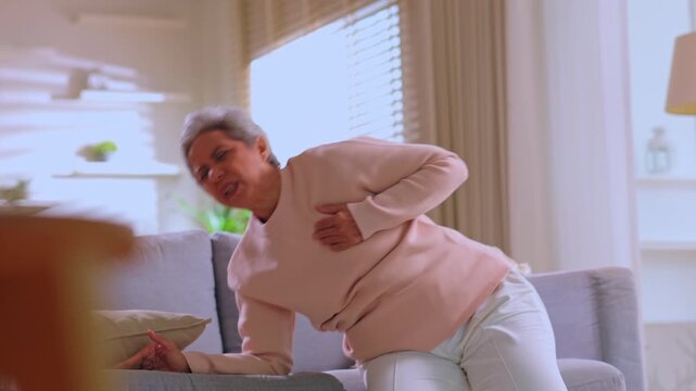 Distressed senior elderly woman suffering sudden chest pain illness indicating potential heart attack during acute medical emergency sitting on living room sofa requiring urgent care