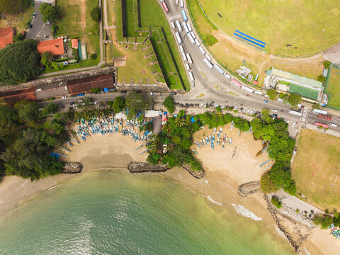 Aerial view of Galle Fort historic walls and colorful fishing boats lined up on the sandy beach with a busy road and lush greenery in Galle, Southern Province, Sri Lanka.