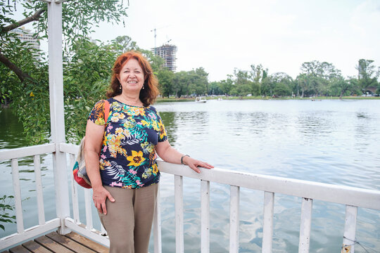 A senior hispanic woman smiles and looks at the camera as she enjoys her free time in a park by a lake. Concept: the well-being of older adults who stay active.