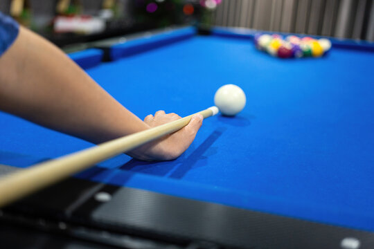Close-up of a person's hand aiming a cue stick at a white cue ball on a blue billiard table, preparing for a shot with colorful pool balls in the blurred background. Sport action scene.