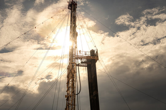 A low-angle perspective of an onshore oil drilling rig structure and derrick tower with cables against a dramatic sunset sky with clouds.