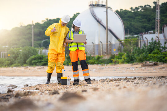Environmental scientists in protective gear testing water quality at industrial site, Professional team performing hazardous material analysis and environmental assessment near oil refinery coastline
