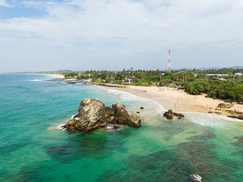 Aerial view of a large rock formation in turquoise water near a sandy beach with palm trees and a communication tower in Koggala, Southern Province, Sri Lanka.