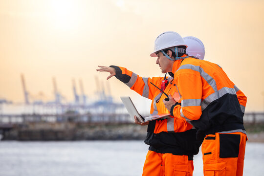 Industrial logistics manager and site supervisor discussing port operations with laptop at sunset, Engineers team planning shipping logistics at commercial harbor terminal