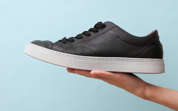 Man's hand holds a black classic leather sneaker with a light sole on a beige background