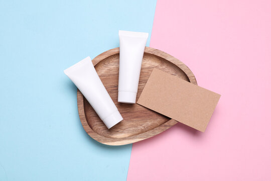 White blank cream tubes with business card in wooden tray on pink blue background. Beauty concept. Top view
