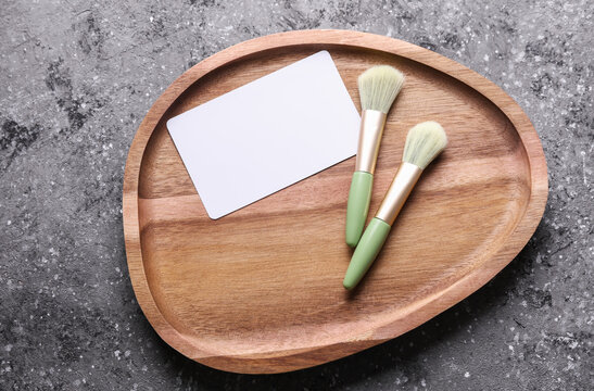 Makeup brushes in wooden tray and white blank business card on concrete background. Beauty concept