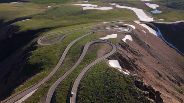 Aerial of Beartooth Highway in Montana USA reveals dramatic serpentine curves through alpine tundra. Drone captures winding mountain pass with scattered snow patches and green meadows on sunny day.