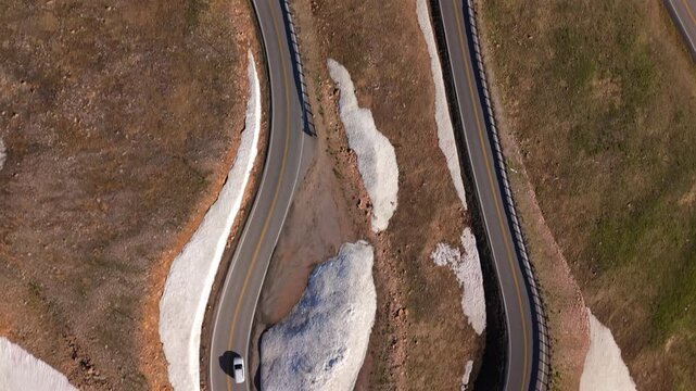 Aerial of Beartooth Highway Montana USA shows drone view of winding mountain road with hairpin turns. Car navigates curved asphalt path through alpine terrain with melting snow patches and green.