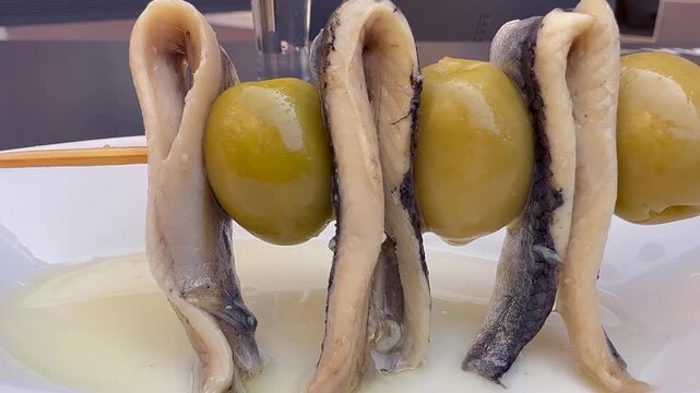 Etreme closeup view of the hand of a man picking a skewer of anchovies and green olives on a white plate with olive oil.