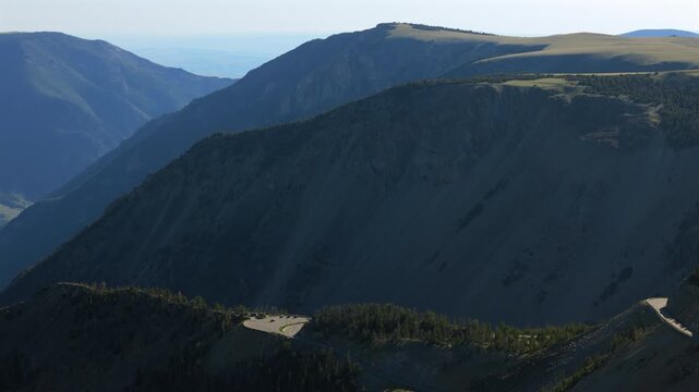 Aerial view of Beartooth Highway winding through Montana mountains with dramatic steep ridges, deep shadowed valleys, scattered evergreen forests, and hazy blue atmosphere under clear summer skies.