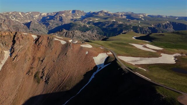 Aerial of Beartooth Highway in Montana, USA showing winding road through alpine terrain with snow patches and green meadows. Drone captures stunning mountain peaks with dramatic cliffs and valleys.
