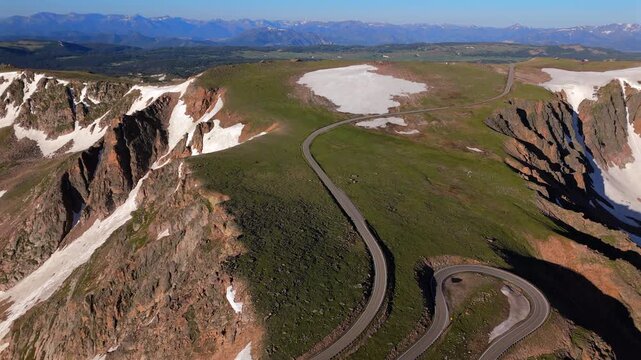 Aerial drone footage of Beartooth Highway winding through dramatic alpine landscape with snow patches dotting green meadows, rugged red rock cliffs, distant mountain ranges under clear blue summer.