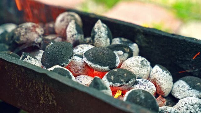 Close up of burning charcoal in metal brazier with glowing embers and ash. Outdoor cooking scene with hot coals, smoke, and heat.