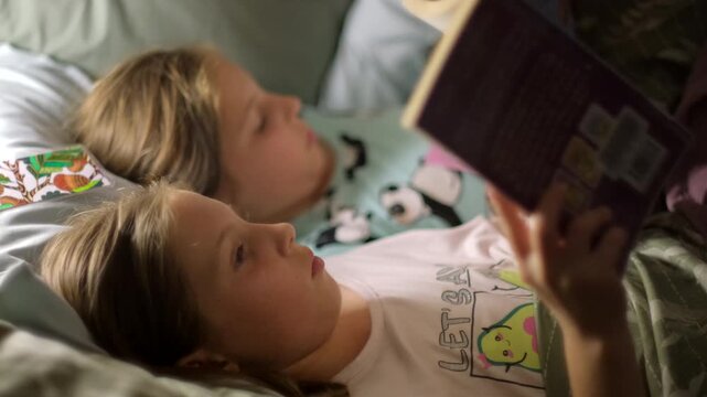 Twin sisters lying in pajamas together in bed reading bedtime stories at night