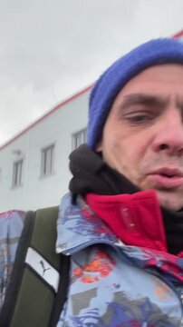 White man vlogging outdoors with beanie, cold urban street setting, colorful patterned jacket and red inner layer, black scarf and backpack visible,