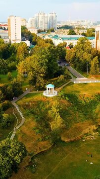 Vertical video. Oryol, Russia. The white rotunda of Dvoryanskoe Gnezdo Park, a classical gazebo perched on a grassy hilltop amid dense summer foliage, aerial descent view toward the pavilion. Drone f