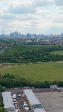 London skyline in distance. Vertical drone reverse over Hackney Marshes, New Spitalfields Market, Eastway and the A12 corridor. Wide aerial view of green fields, roads and city.