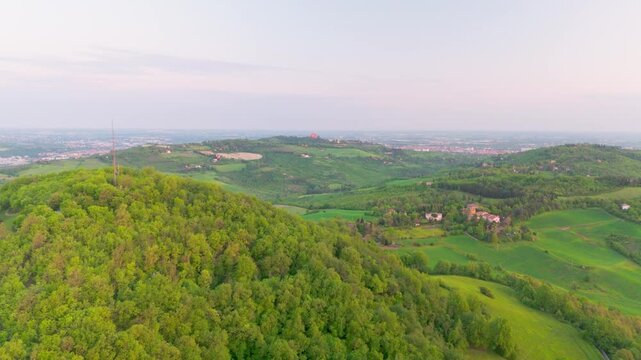 Drone lifting at dawn over Parco di Forte Bandiera on Monte Donato, revealing green forested hills and the distant city of Bologna in soft morning light.