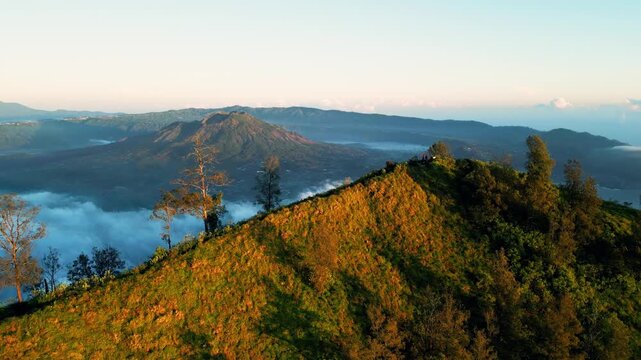 Mount Batur volcano and crater lake with green mountain slopes in golden morning light across high altitude terrain in Bali with sea of clouds smooth aerial drone shot