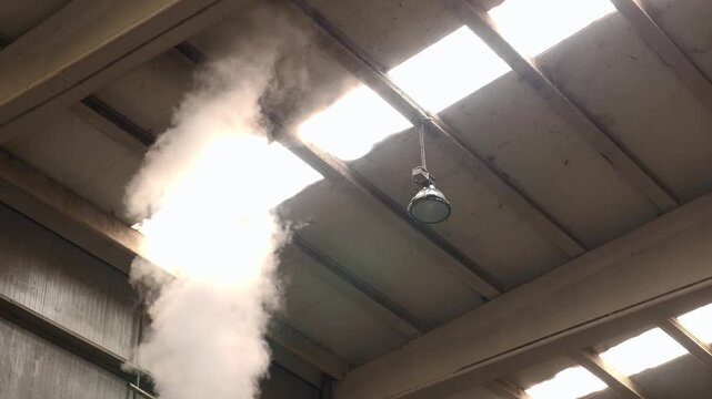 Low angle static shot of thick white steam rising and venting out through a skylight on a factory ceiling next to an industrial lamp.