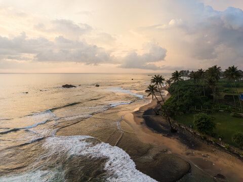 Aerial view of Gintota beach with palm trees lining the sandy shore and waves crashing against the coast under a cloudy sunset sky in Galle, Southern Province, Sri Lanka.