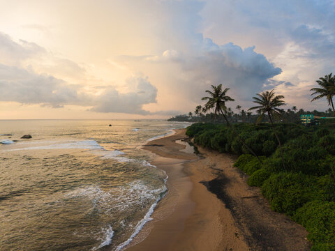 Aerial view of Gintota beach with palm trees, lush greenery, and waves crashing on the sand under a dramatic sunset sky in Galle, Southern Province, Sri Lanka.