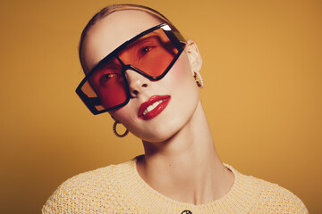 Stylish woman with short hair wearing oversized red sunglasses and gold hoop earrings poses against a warm yellow background. Fashion concept with bold accessories and makeup. © SHOTPRIME STUDIO