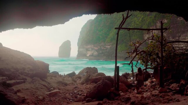 Cinematic view of woman on wooden swing inside a hidden beach cave in Nusa Penida, Bali 4K