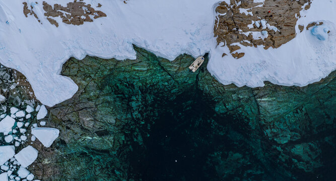 Aerial view of a small boat moored at the edge of a snowy shoreline with turquoise arctic waters and scattered ice floes Tasiilaq, Sermersooq Municipality, Greenland.