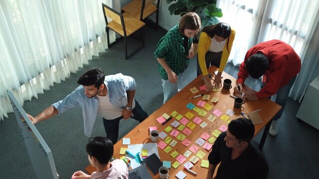 Top view of diverse business people writing and stick sticky notes at whiteboard. Creative team working together and brainstorming marketing idea for startup project at table with notes. Convocation.