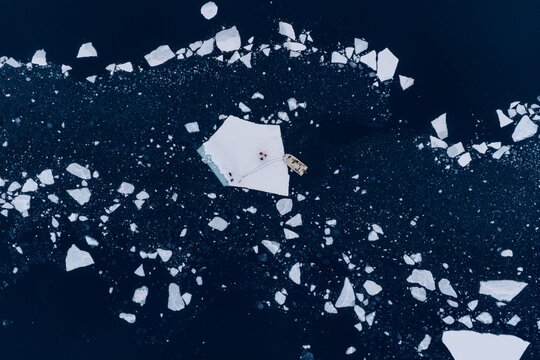Aerial view of a small boat moored to a large flat iceberg with people standing on it in the dark arctic waters Tasiilaq, Sermersooq Municipality, Greenland.