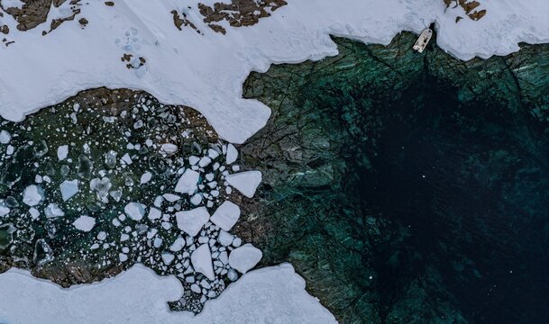 Aerial view of a small boat moored by a snow-covered rocky shoreline with turquoise water and floating ice floes in Tasiilaq, Sermersooq Municipality, Greenland.