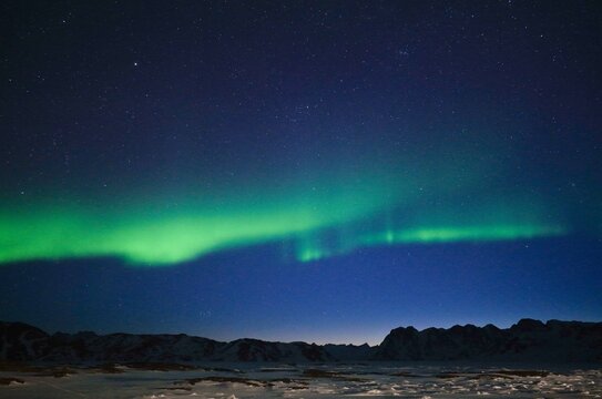 Aerial view of the vibrant green Aurora Borealis dancing across a starry night sky over snow-covered mountains in Tasiilaq, Sermersooq Municipality, Greenland.